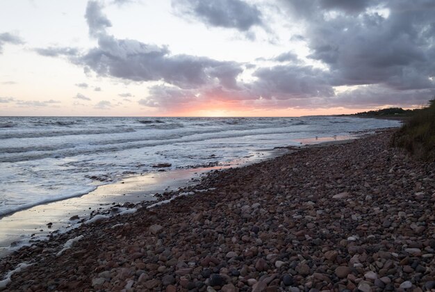 Foto puesta de sol en una playa de guijarros con el sol en el horizonte y bajo un cielo parcialmente nublado