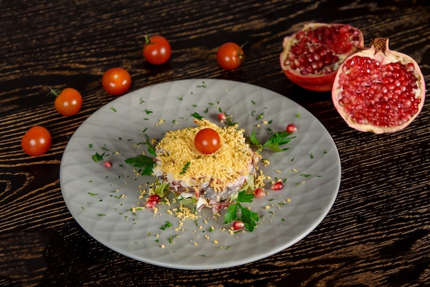 Puff Salat mit Fleisch, Gemüse, Käse, Eiern, garniert mit Kräutern und Granatapfel und Kirschtomaten auf einem grauen Teller. Vor dem Hintergrund von Granatapfel- und Kirschtomatenhälften.
