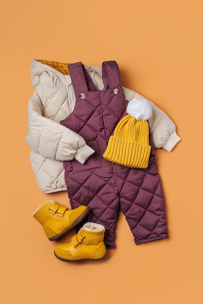 Pufferjacke, warme Hose, Mütze und Stiefel auf orangem Hintergrund. Set Babykleidung für den Winter. Mode Kinder Outfit.