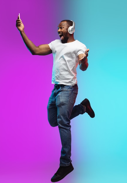 Pulando alto. Retrato de jovem afro-americano em fundo gradiente em neon. Bela modelo masculino em estilo casual, camisa branca. Conceito de emoções humanas, expressão facial, vendas, anúncio.