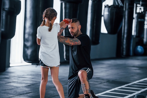 Puñetazos básicos. Entrenador de boxeo tatuado joven enseña a niña linda en el gimnasio.