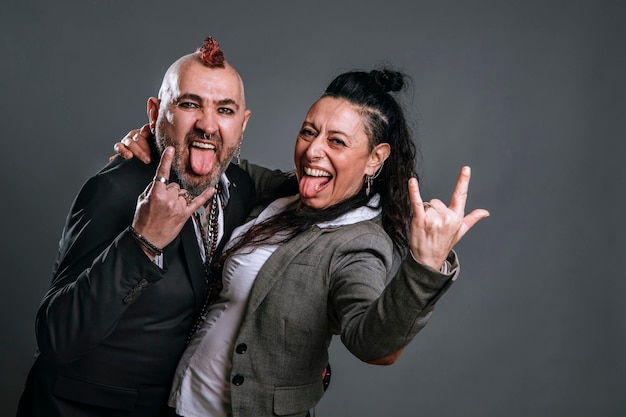 Punkstyle-Mann und -Frau, die Jackenanzüge tragen, posieren in Studioaufnahmen und schauen in die Kamera, die ihre Zungen herausstreckt und mit fröhlichem und lässigem Ausdruck Hörner mit den Händen macht