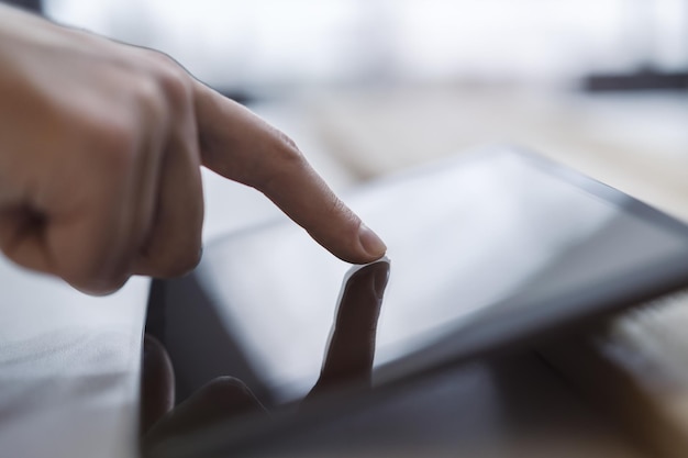 La punta del dedo del hombre tocando suavemente la pantalla de una tableta digital moderna tendida en un escritorio rodeado por el brillo desenfocado de un espacio de oficina contemporáneo