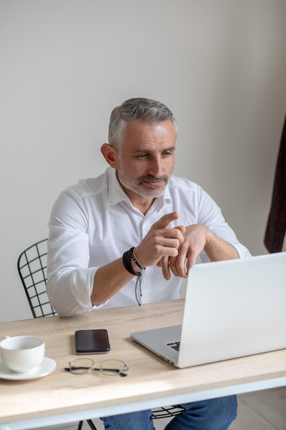 Punto importante. Hombre de mediana edad de pelo gris de negocios mirando el portátil señalando con el dedo a la pantalla sentado en la mesa en una habitación luminosa