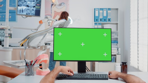 Punto de vista del asistente que trabaja con pantalla verde en la computadora en el consultorio del dentista. Hombre que usa el monitor con clave de croma y una plantilla de maqueta aislada para odontología y cuidado de los dientes en el gabinete dental
