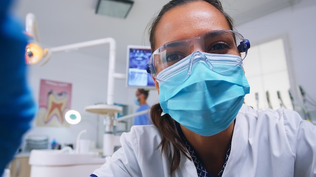 Punto de vista del dentista que trabaja en la higiene de la boca del paciente en el consultorio dental, comprobando los problemas de los dientes. Médico examina en consultorio de ortodoncia con lámpara de luz y utensilios esterilizados, cara de primer plano en máscara médica.