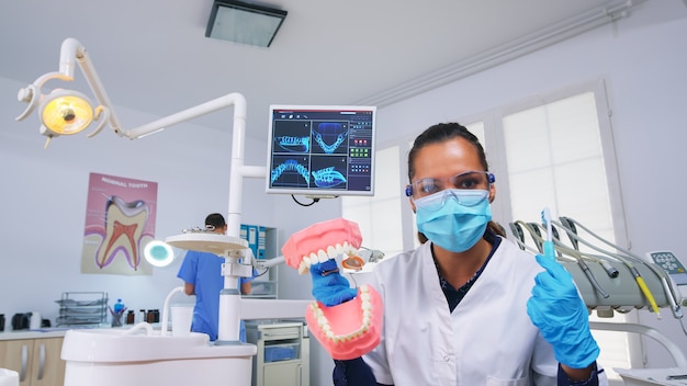 Punto de vista del paciente del dentista que enseña a la mujer la limpieza correcta de los dientes en el consultorio dental con el accesorio de esqueleto. Estomatólogo con máscara de protección pidiendo presentación objeto médico de los dientes