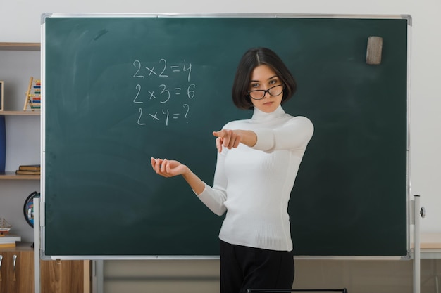 puntos estrictos al lado joven maestra con anteojos de pie frente a la pizarra y escribe en el aula