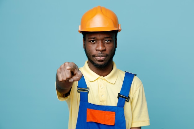 Puntos estrictos en la cámara joven constructor afroamericano en uniforme aislado sobre fondo azul.