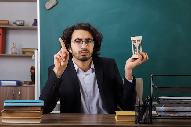 Puntos impresionados en el profesor de sexo masculino con gafas sosteniendo un reloj de arena sentado a la mesa con herramientas escolares en el aula