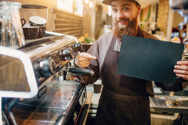 Puntos de joven emocionado en un trozo de papel oscuro. Lo sostiene en la mano y mira a la cámara. Guy de pie en la máquina de café en la cocina.