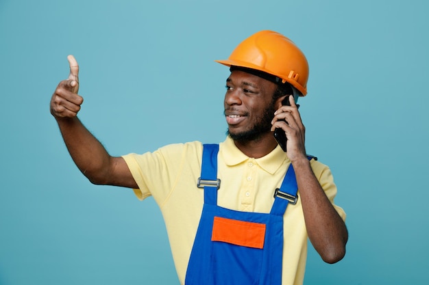 Puntos sonrientes al lado joven constructor afroamericano en uniforme habla por teléfono aislado sobre fondo azul.