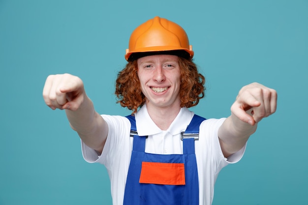 Puntos sonrientes en cámara joven constructor hombre en uniforme aislado sobre fondo azul.