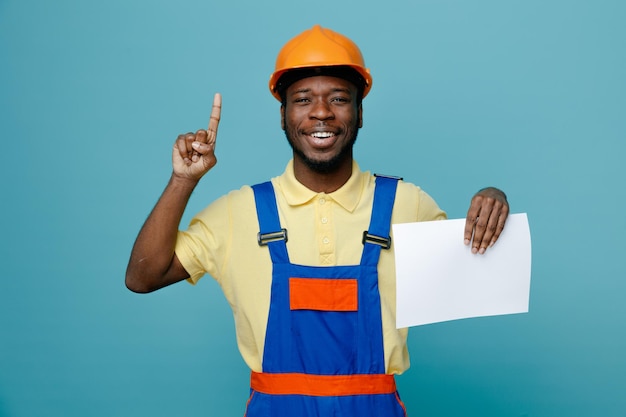 Puntos sonrientes en un joven constructor afroamericano en papel de sujeción uniforme aislado en fondo azul