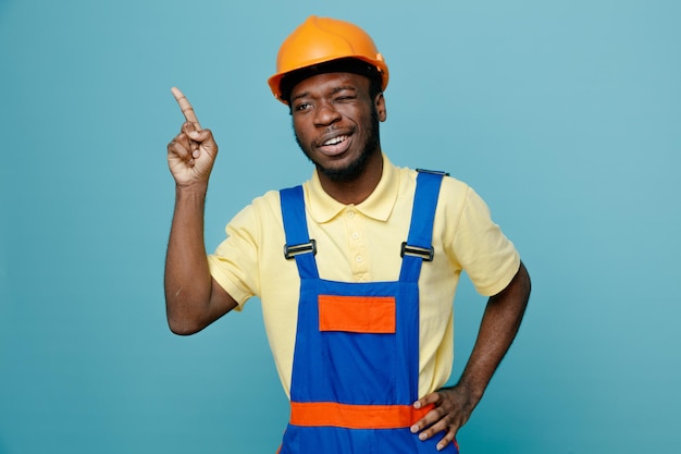 puntos sonrientes en poner la mano en las caderas joven constructor afroamericano en uniforme aislado sobre fondo azul