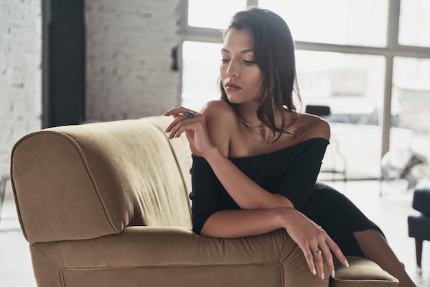 Pura elegancia. Atractiva mujer joven en elegante vestido negro simplemente descansando mientras está sentado en el sofá
