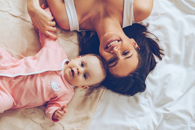 Pura felicidad. Vista superior de la alegre hermosa joven mirando a la cámara con una sonrisa mientras está acostado en la cama con su bebé