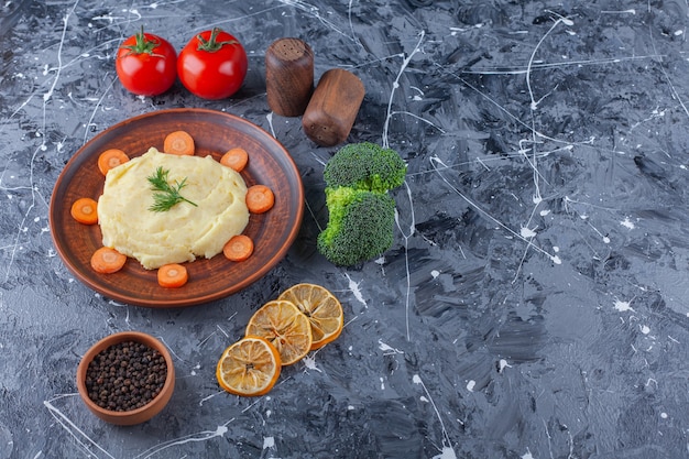 Purê de batata e cenouras fatiadas em um prato ao lado de legumes e tigelas de temperos, sobre o fundo azul.