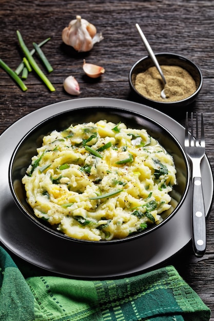 Purê de batata e couve - acompanhamento irlandês chamado colcannon com alho, manteiga derretida, pimenta preta por cima, servido em um prato preto com talheres em um fundo de madeira escuro, vista de cima, close-up