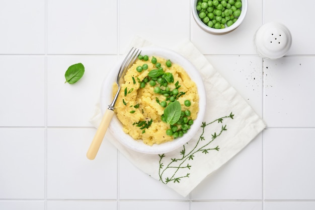 Puré de patatas con mantequilla, guisantes, cebollas, albahaca sobre fondo de baldosas de cerámica blanca.