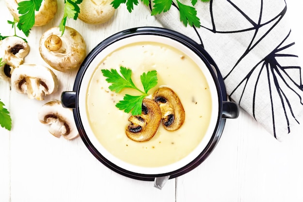 Puré de sopa de champiñones, papas, cebollas y crema en un tazón, toalla de cocina, perejil y cuchara sobre fondo de tablero de madera desde arriba