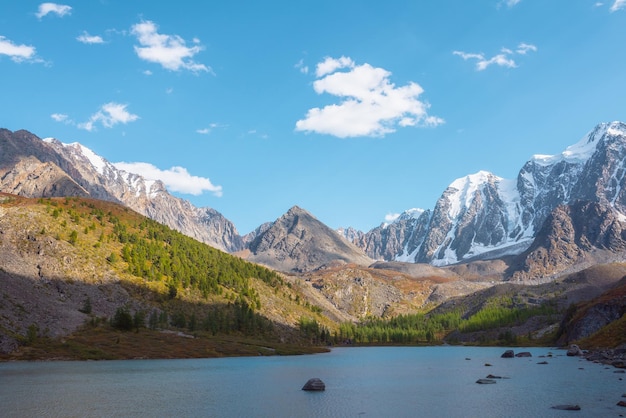 Puro lago alpino azul con vista a la colina del bosque y las montañas gigantes de nieve en el día soleado de otoño Lago glacial contra una enorme cordillera cubierta de nieve al sol brillante Colores vívidos de otoño en las altas montañas