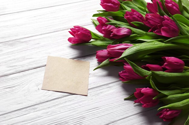 Púrpura hermosos tulipanes con tarjeta de felicitación sobre fondo blanco de madera