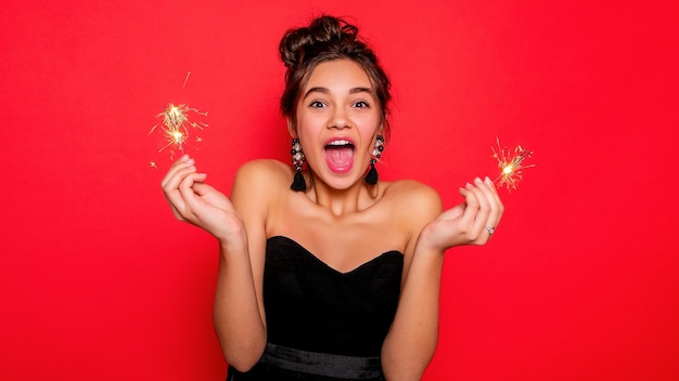 Pyrotechnik und People-Konzept Lächelnde junge Frau oder Teenager-Mädchen glückliche Frau mit Wunderkerzen feiern in schwarzem Kleid auf rotem Hintergrund glücklich mit offenem Mund