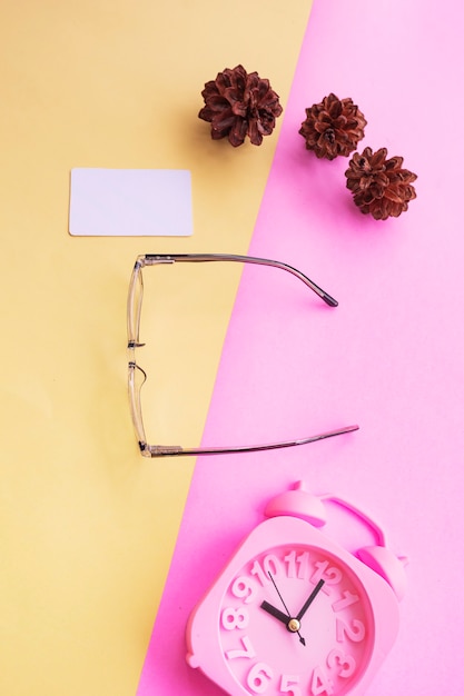 Quadratische Brille auf dem Foto im minimalen Sommerstil auf pastellrosa und gelbem Hintergrund. Wecker , Kiefernblüten , Visitenkarten