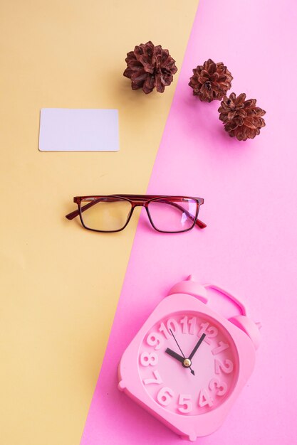 Quadratische Brille auf dem Foto im minimalen Sommerstil auf pastellrosa und gelbem Hintergrund. Wecker , Kiefernblüten , Visitenkarten