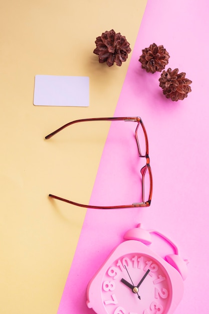 Quadratische Brille auf dem Foto im minimalen Sommerstil auf pastellrosa und gelbem Hintergrund. Wecker , Kiefernblüten , Visitenkarten