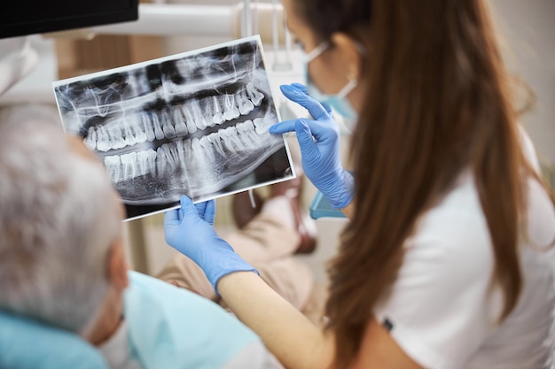 Qualifizierte junge Zahnärztin, die ein Röntgenbild der Zähne hält und es ihrem älteren Patienten zeigt