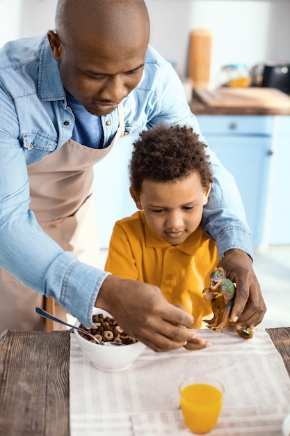 Qualitätszeit zusammen. Charmanter junger Vater, der mit einem Spielzeugdinosaurier zusammen mit seinem Sohn spielt und das Spielzeug mit Müsli füttert