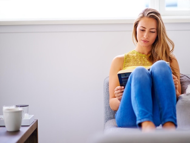 Quando ela está lendo, ela está relaxando Foto de uma jovem lendo um livro em seu sofá em casa