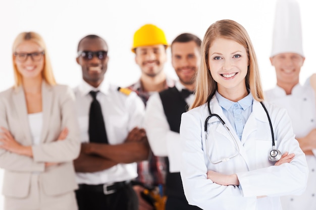 Foto quando eu crescer, serei médico. bela jovem médica de braços cruzados e sorrindo enquanto um grupo de pessoas em diferentes profissões em pé no fundo