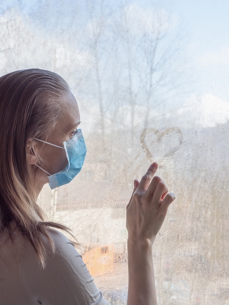 Quarantäne, Coronavirus-Pandemie. Eine Frau trägt eine medizinische Einwegmaske, steht traurig am Fenster und zeichnet ein Herz auf das nasse Glas