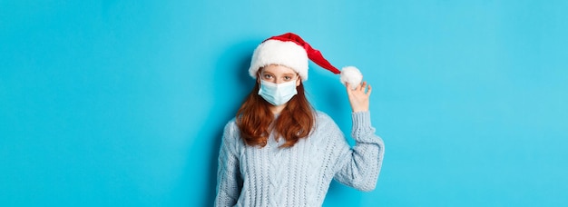 Foto quarentena de natal e ruiva de conceito cobiçoso usando máscara facial e brincando com cel de chapéu de papai noel