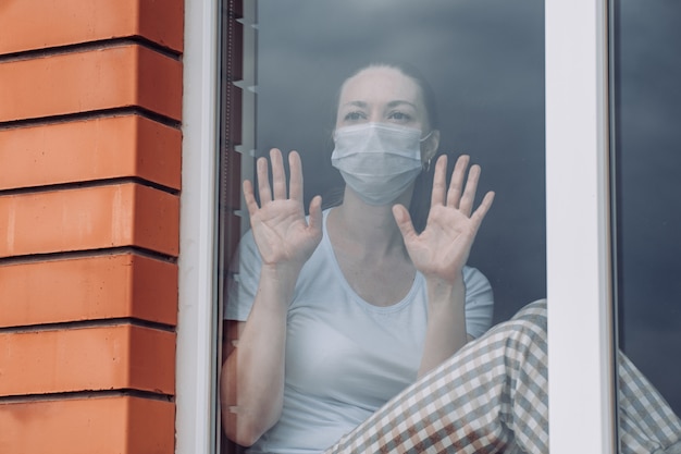 Foto quarentena em casa. mulher caucasiana, sentado na janela em uma máscara médica, olhando para fora, quer sair. proteção contra infecção por coronavírus.