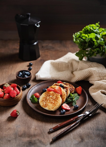 Quarkpfannkuchen oder Syrniki mit Minze, Erdbeeren und Heidelbeeren auf einem dunklen Holztisch mit schwarzer Kaffeekanne. Konzept dunkles und launisches gesundes Frühstück.