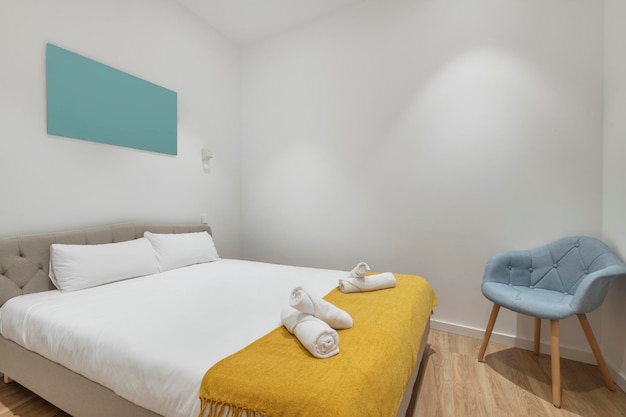 Quarto com cama king size com edredom branco pequeno cobertor amarelo toalhas enroladas na cama tecido capitone azul cadeira estofada em apartamento de aluguel por temporada