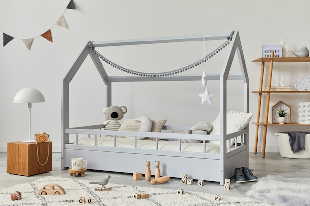 Quarto infantil escandinavo elegante com cama de madeira criativa, cubo de madeira, abajur, prateleira de madeira, brinquedos de pelúcia e madeira e decorações têxteis penduradas. Paredes cinzentas. Modelo.