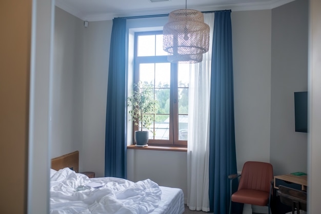 Quarto, interior. Cama e janela com cortinas e planta de casa em um pequeno quarto aconchegante em estilo minimalista com luz natural