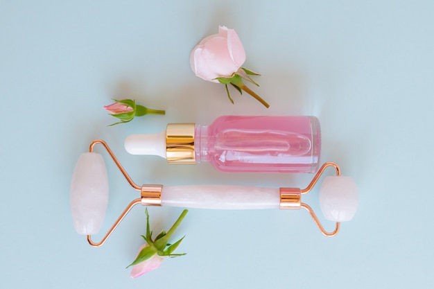 Quartz Roller Schönheitsölprodukte auf blauem Hintergrund mit Rosenknospen. Serum oder Öl und Hautpflegetools. Wellness-Konzept. Ansicht von oben.