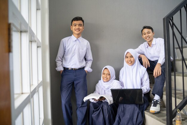 Quatro adolescentes com uniformes do ensino fundamental sorriem para a câmera enquanto seguram um laptop e um livro