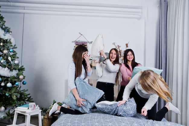 Quatro amigas fofas usam suéteres quentes e calças pretas na cama no quarto decorado de ano novo no estúdio, brincam com travesseiros.