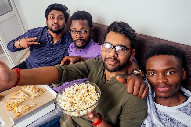 Quatro amigos afro-americanos fazendo selfie na câmera do smartphone com pizza italiana e pipoca na festa em casa