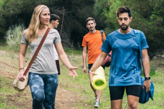 Quatro amigos conversando relaxados enquanto carregam esteiras e bolsas de ioga para passear na floresta