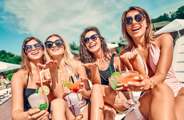 Quatro belas mulheres curtindo suas férias de verão com coquetéis à beira da piscina Vista de perto