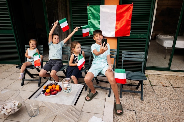 Quatro crianças felizes com bandeiras italianas comemorando o Dia da República da Itália Garoto fazendo selfie no telefone