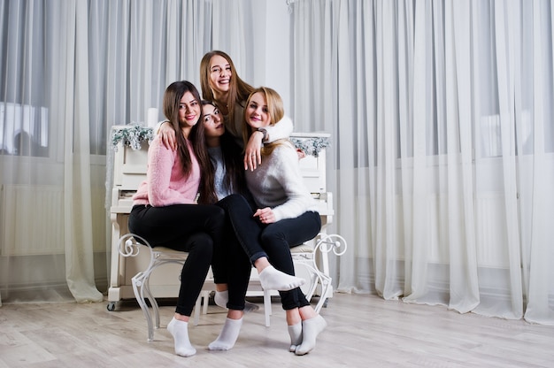 Quatro garotas de amigos fofos usam suéteres quentes e calças pretas contra o velho piano com decoração de Natal no quarto branco.